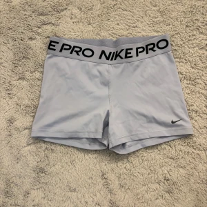 Nike Pro ljusgrå träningstights-shorts - Nike Pro shorts i ljusgrått med bred resår i midjan där det står NIKE PRO i svart. Tight passform och korta ben, perfekta för träning eller sport. Snygg och sportig look med diskret Nike-logga framtill. Gjorda i stretchigt syntetmaterial för maximal rörelsefrihet.