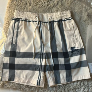 Burberry rutiga shorts med snörning - Snygga Burberry shorts i klassiskt beige med mörkblå och grå rutor. De har elastisk midja med vit snörning och sidofickor. Ikoniskt Burberry-tryck på ena benet. Perfekta för sommaren och ger en clean, lyxig vibe.