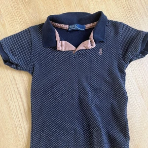 Mörkblå pikétröja från Polo Ralph Lauren - Snygg mörkblå pikétröja från Polo Ralph Lauren med diskret rutmönster och klassisk krage i kontrastfärg. Tröjan har korta ärmar och broderad logga på bröstet. Perfekt för en stilren och avslappnad look. I storlek 122-128 alltså till barn 
