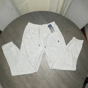 Grå mjukisbyxor Polo Ralph Lauren - Ljusgrå mjukisbyxor från Polo Ralph Lauren med elastisk midja och snörning. Byxorna har ribbade muddar vid bensluten och en liten broderad logga på ena benet. Tillverkade i mjukt bomullsmaterial för maximal komfort.