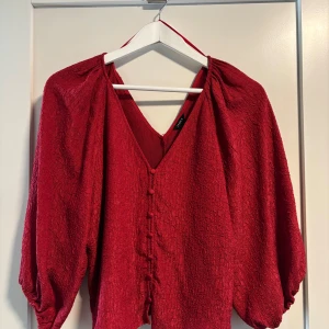 Röd blus med v-ringning och knappar - Säljer en röd blus i storlek M från Lindex med v-ringning både fram och bak. Blusen har ballongärmar, täckta knappar framtill 
