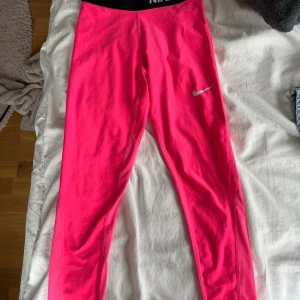 Nike Pro rosa  - Snygga och sportiga Nike Pro tights i starkt rosa med svart resår och vit logga. Tight passform och stretchigt material som andas, perfekta för träning eller gym. Midjan är markerad med bred svart resår där det står NIKE PRO.