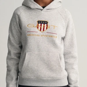 Grå hoodie från GANT med tryck - Snygg ljusgrå hoodie från GANT med broderat logotyp-tryck och sköld på bröstet. Tröjan har huva, känguruficka och ribbade muddar. Perfekt för en chill och sportig look. Tillverkad i mjuk bomullsblandning för extra komfort. Köpt för 900 kr men säljer för 250kr. Tröjan har inga defekter förutom initialer på tvätt lappen, dessa kan dock klippas bort enkelt 