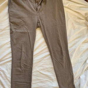 Beige kostymbyxor från J.Lindeberg - Snygga beige kostymbyxor från J.Lindeberg i klassisk rak modell. Byxorna har fickor fram och bak med knappdetaljer, samt stängs med knapp och dragkedja. Perfekta för en clean och stilren look. 