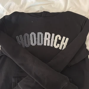 Svart HOODRICH hoodie med tryck - Svart hoodie från HOODRICH med stort broderat logotyp-tryck i grått och vitt över bröstet. Tröjan har huva med snörning, känguruficka och dragkedja på ena ärmen. Perfekt för dig som gillar streetwear och vill ha en bekväm och snygg look. Tröjan saknar snören men detta påverkar inte tröjans användning. Tröjan har fickor vid armarna vilket är perfekt för att säkra grejer.