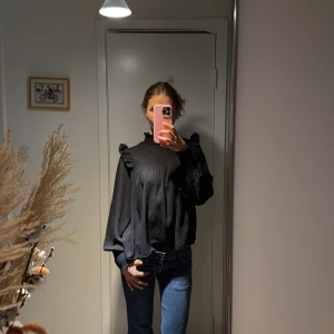 Svart volangblus med smockad krage - Svart blus med högt smockat parti vid halsen och axlarna, volangdetaljer och långa puffiga ärmar med smockade muddar. Luftig och flowy passform, perfekt till jeans eller kjol. Tidlös och stilren design som ger en cool vibe.