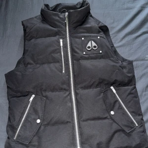 Svart dunväst från Moose Knuckles - Snygg svart äkta  dunväst från Moose Knuckles med hög krage, silverfärgade dragkedjor och logga på bröstet. Västen har två fickor med dragkedja och tryckknapp samt broderad detalj på insidan. 