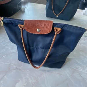 Marinblå Longchamp handväska - Säljer en klassisk marinblå handväska från Longchamp med bruna skinnhandtag och lock. Väskan har dragkedja upptill och är tillverkad i slitstark syntet med detaljer i skinn. Perfekt storlek för vardagens alla prylar och enkel att matcha till olika outfits. 