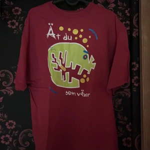 Röd Hanes t-shirt med grafiskt tryck - Cool röd t-shirt från Hanes med ett stort grafiskt tryck framtill i grönt, gult och blått. Texten 'Ät du som växer' och en unik illustration gör den till ett riktigt statement-plagg. Skön passform och tillverkad i 100% bomull.