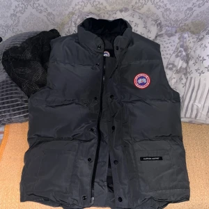 Svart dunväst från Canada Goose - Säljer en svart dunväst från Canada Goose med klassisk logga på bröstet. Västen har hög krage, dragkedja och tryckknappar framtill samt två stora fickor. Perfekt för lager-på-lager och riktigt snygg till streetwear.