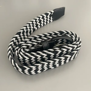 Svartvitt flätat bälte i textil - Snyggt svartvitt bälte med flätad design och svarta skinn-detaljer vid spännet. Bältet har ett coolt randigt mönster och är gjort i slitstark textil som ger en modern och sportig vibe. Perfekt för att lyfta vilken outfit som helst med lite extra edge.