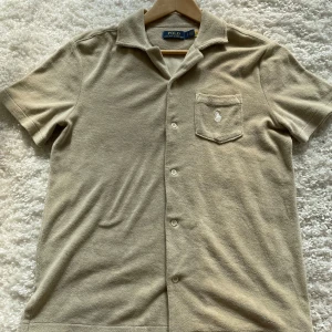 Beige Frotté pike Polo Ralph Lauren - Snygg beige skjorta från Polo Ralph Lauren i frotté. Pikén har korta ärmar, klassisk krage, knäppning framtill och en bröstficka med broderad logga. 