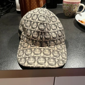 Mönstrad keps från Salvatore Ferragamo - Snygg keps från Salvatore Ferragamo med ikoniskt grått och beige mönster över hela kepsen. Klassisk böjd skärm och justerbar baksida. Perfekt accessoar för att lyfta din outfit med exklusiv känsla.