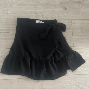 Svart omlottkjol från NA-KD x Pamela - Supersnygg svart kort omlottkjol från NA-KD x Pamela med volangkant och knyt i midjan.