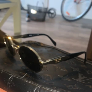 Prada solglasögon  - Snygga solglasögon från Prada med rund form och svarta glas. Bågarna är i guldmetall med svarta skalmar och diskret Prada-logga på sidan. Perfekt accessoar för en trendig look och passar dig som gillar klassisk men ändå modern stil.