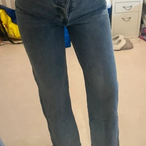 Blå raka jeans med hög midja - Säljer ett par klassiska blå jeans med hög midja och raka ben. Jeansen har en enkel design med två bakfickor och stängs med dragkedja och knapp. Perfekta för en chill och stilren look.