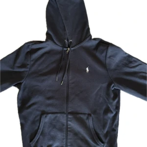 Svart hoodie från Polo Ralph Lauren - Svart hoodie från Polo Ralph Lauren med klassisk broderad logga på bröstet. Tröjan har dragkedja, huva med snörning och två stora fickor framtill. Tillverkad i mjuk bomullsmix, perfekt för en chill och stilren look.