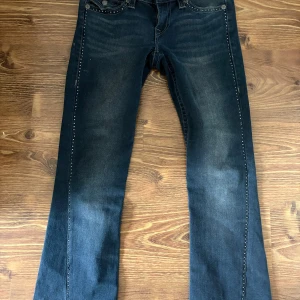 True Religion Joey Low Rise Flare Jeans - Mörkblå jeans från True Religion, modell Joey Low Rise Flare. Snygga kontrastsömmar, ficklock med knapp bak och coola nitar längs sidorna. Bootcut/flare passform med låg midja. Klassisk denim med slitningar för en trendig look.