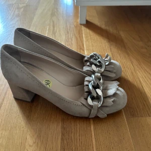 Beige mocka pumps från Rizzo - Vi kan komma överens om priset! Stilrena pumps i beige mocka från Rizzo med chunky klack och rund tå. Skorna har dekorativa fransar och en stor silverfärgad kedjedetalj framtill som ger en cool touch. Perfekta för dig som gillar klassisk stil med en twist.