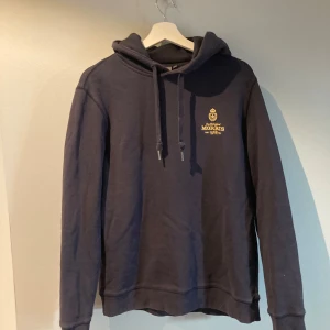 Mörkblå hoodie från Morris, storlek M - Snygg mörkblå hoodie från Morris med broderad logga och text på bröstet. Tröjan har huva med snörning och är tillverkad i mjuk bomull. Nypriset ligger på 1799kr. 