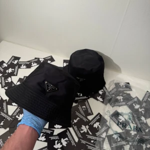 Svart bucket hat från Prada - Stilren svart bucket hat från Prada med ikonisk triangellogo i metall framtill. Hatten har klassisk form med bred, nedåtlutande brätte och är tillverkad i glansig syntet. Perfekt accessoar för en trendig look.