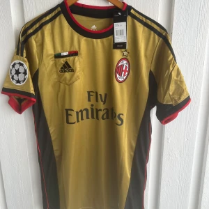 AC Milan Kaka' Champions League tröja - Grym AC Milan fotbollströja från Adidas i guld med svarta och röda detaljer. Champions League-märke på ärmarna, klubbmärke och Fly Emirates-tryck på bröstet. Kaka' och nummer 22 på ryggen. Tillverkad i lätt och glansigt material, perfekt för fans.
