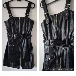 Svart kort klänning i skinnimitation - Cool svart kort klänning i skinnimitation med breda axelband, dragkedja framtill och bälte i midjan. Snygga detaljer som ficka med knapp och silvriga spännen. Perfekt för dig som vill sticka ut med en edgy look.