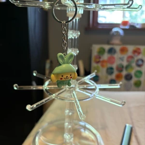 Söt nyckelring med kaninfigur - Nyckelring med en gullig figur i form av en gul hund med gröna kaninöron och gröna kläder. Figurerna har röda kinder och ett glatt ansikte, perfekt att hänga på väskan eller nycklarna för en extra söt detalj.