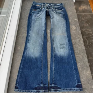Blå bootcut jeans från G-Glas - Snygga blå bootcut jeans från G-Glas med slitna detaljer, patchade partier och fransade kanter vid fickor och midja. Jeansen har låg midja, dubbla knappar och klassisk femficksdesign. Perfekta för dig som gillar en cool och avslappnad stil. Midjemått rak över är 37,5cm och längden är 80cm