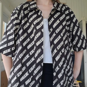 Svart Billie Eilish skjorta med tryck - Cool svart skjorta med vitt Billie Eilish-tryck över hela plagget. Oversized passform, kortärmad och knäppning framtill. Officiell kollektion från H&M. Använd endast en gång.