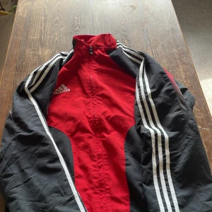 Röd och svart Adidas vindjacka M - Säljer en sportig röd och svart vindjacka från Adidas med klassiska vita ränder längs ärmarna och dragkedja framtill. Jackan har hög krage och meshfoder på insidan för extra komfort. Perfekt för dig som gillar streetwear och sportig stil.