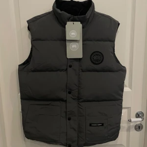 Mörkgrå dunväst från Canada Goose - Säljer en mörkgrå dunväst från Canada Goose med hög krage, två stora fickor framtill och diskret svart logotyp på bröstet. Västen har knappar och dragkedja framtill och är perfekt för lager-på-lager-stil under kyliga dagar. Inte äkta.