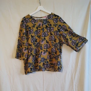 Blommig blus från Indiska i senapsgult - Blus från Indiska i senapsgult med blommigt mönster i vit, orange, blått och svart. Modellen är loose fit med trekvartsärm och rund halsringning. Perfekt för dig som gillar färg och mönster!