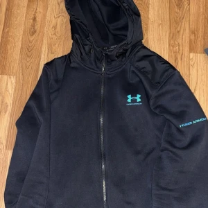 Svart vindjacka från Under Armour - Svart vindjacka från Under Armour med huva och hel dragkedja. Jackan har blå logga på bröstet och ärmen, samt paneler i blankare material över axlarna. Perfekt för dig som gillar sportig stil och vill ha något lätt och smidigt.