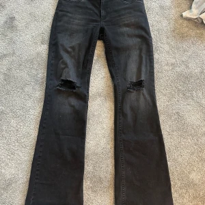 Svarta bootcut jeans från Nelly - Snygga svarta bootcut jeans från Nelly med låg midja och slitna hål på båda knäna. Jeansen har klassisk femficksdesign och är tillverkade i stretchigt denim för en bekväm passform. Perfekta för en cool och avslappnad stil. Använda en gång så dem är som nya men utan prislapp. 