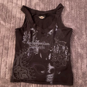 Svart Harley Davidson linnetopp med tryck - Cool svart linnetopp från Ed Hardy med grått paisley- och blommönster samt glittriga detaljer framtill. V-ringad och ärmlös modell i mjuk bomull, perfekt för dig som gillar edgy streetstyle.