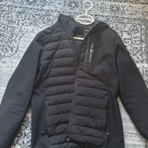 Svart pufferjacka från Sail Racing - Säljer en svart pufferjacka från Sail Racing med huva och dragkedja. Jackan har quiltade partier och är tillverkad i syntetmaterial med sportig look. Perfekt för kyliga dagar och har en clean design med logga på bröstet och ärmen. Kan diskutera pris