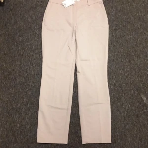 Beige kostymbyxor från H&M, stl 40 - Snygga beige kostymbyxor från H&M i rak modell. Byxorna har hällor för bälte, diskreta fickor och stängs med hake och dragkedja. Perfekta för en clean och stilren look. Materialet är mjukt och känns bekvämt hela dagen.