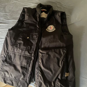 Svart dunväst från Moncler - Säljer en svart dunväst från Moncler den heter Bourges Gilet 1952 med klassisk logga på bröstet. Västen har flera stora fickor framtill, dragkedja och hög krage. Den är quiltad och har en clean, sportig look som funkar till många outfits. Nypris är 7000kr. Vid frågor är det bara att skriva. 