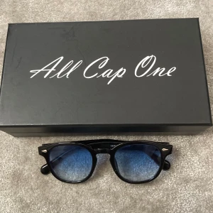Svarta All Cap One solglasögon - Snygga svarta solglasögon från All Cap One med klassisk rundad form och blåtonade glas. Bågarna är i blank svart plast med silvriga detaljer vid gångjärnen. Perfekta för att lyfta din stil med en cool touch.