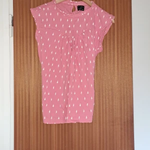 Rosa blus Orla Kiely x People Tree - Supersöt rosa blus med vita figurmönster från Orla Kiely x People Tree. Blusen har korta ärmar, rund hals med liten knapp bak och knyt i ryggen för extra passform. Tillverkad i mjuk bomull, perfekt för vår och sommar.