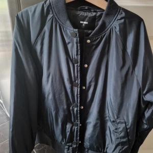 Svart bomberjacka från Bik Bok - Snygg svart bomberjacka från Bik Bok med klassisk ribbad krage och knappar framtill. Jackan har lång ärm och två fickor på sidorna. Perfekt för dig som vill ha en stilren och enkel jacka till vardagslooken.