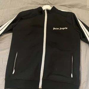 Palm Angels svart track jacket M - Svart track jacket från Palm Angels med vita ränder längs ärmarna och vit dragkedja. Klassisk logga på bröstet och två fickor med dragkedja. Jackan är i sportig stil och tillverkad i bomull och polyester för en skön passform.