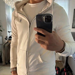 Vit hoodie från Polo Club med huva - Snygg vit hoodie från Polo Club med dragkedja, huva med snörning och känguruficka. Tröjan har en liten blå logga broderad på bröstet och är gjord i mjuk bomull. Perfekt för en chill och stilren look.