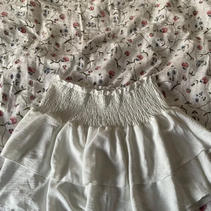 Vit volangkjol med smock - Supersöt vit kortkjol med bred smock i midjan och volanger. Kjolen har en luftig och flowy känsla, perfekt för varma dagar. Passar dig som gillar en feminin och trendig stil.