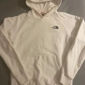 Vit hoodie från The North Face - Snygg vit hoodie från The North Face med stor svart logga och texten 'Never Stop Exploring' på ryggen. Klassisk känguruficka framtill, huva och långa ärmar. Perfekt för dig som gillar streetwear och enkel stil.