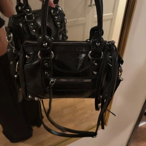 Svart skinnhandväska med dragkedjor citybag - Cool svart handväska i skinn med silvriga metalldetaljer och dragkedjor. Väskan har både handtag och axelrem, samt dekorativa remmar och nitar. Perfekt storlek för det viktigaste och ger en edgy vibe till din outfit.