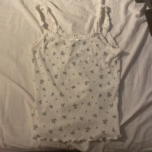 Vit blommig linnetopp från Divided - Supersöt vit linnetopp från Divided med små blå blommor och ribbad struktur. Toppen har spetsdetaljer längs axelbanden och halsringningen samt vågig kant nertill. Perfekt för en somrig och chill look.