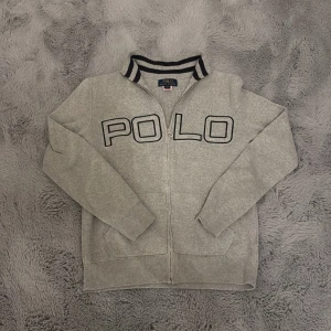 Grå kofta från Polo Ralph Lauren - Snygg grå kofta från Polo Ralph Lauren med dragkedja och stor POLO-logga broderad framtill. Ribbad krage med svarta och vita ränder ger en sportig vibe. Perfekt till jeans eller joggers för en chill look. Tröjan har inga skador.