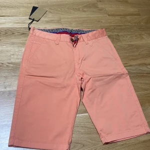 Persikofärgade shorts från Veslor - Snygga persikofärgade shorts från Veslor med klassisk passform och bälteshällor. Tillverkade i mjuk bomull med lite stretch för extra komfort. Flera fickor både fram och bak samt coolt mönstrat innerfoder i midjan. Perfekta för varma dagar!